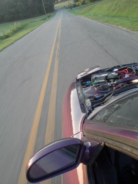 1995 Ford Probe - A car with its hood open is driving down a two-lane road. The driver's side mirror and part of the engine are visible.