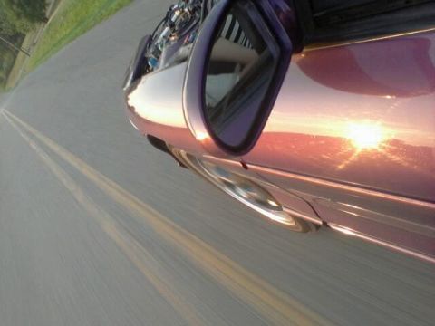 1995 Ford Probe - A low-angle shot of the side of a purple car, reflecting the sunset on its body. The car is moving on a road with double yellow lines.