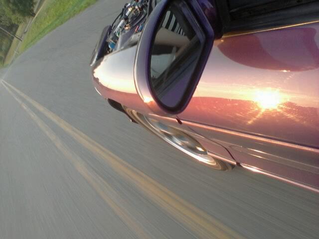 Purple car side view reflecting sunset, driving on a road.