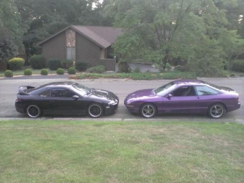 1995 Ford Probe - A black two-door coupe and a purple two-door coupe are parked side-by-side on a paved surface in front of a house.