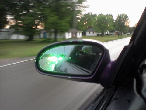 1995 Ford Probe - A car's side mirror reflects bright green lights from another vehicle driving on a road next to a grassy area with trees and houses. The im…