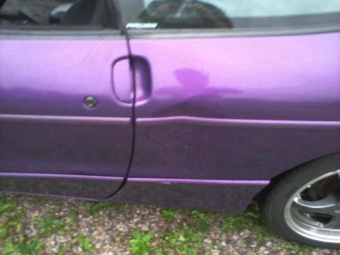 1995 Ford Probe - A close-up side view of a purple car door with a visible dent and a chrome door handle. The car is parked on a gravel and grass surface.