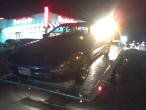 1995 Ford Probe - A dark coupe is loaded onto a tow truck at night, with a brightly lit building in the background.