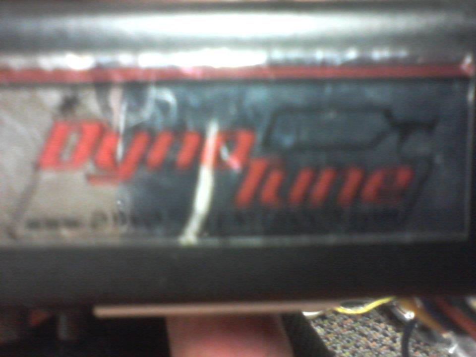 Close-up of a red "DynoTune" sticker on a dark surface, with a Supermotors logo at the bottom right