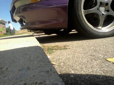 1995 Ford Probe - Low-angle shot of the front bumper and wheel of a purple car parked on asphalt next to a concrete curb. A person in a white shirt and dark…