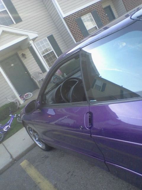 1995 Ford Probe - A low-angle shot of the driver's side of a purple 1995 Ford Probe. The car is parked on asphalt next to a curb, with a building and a bicyc…