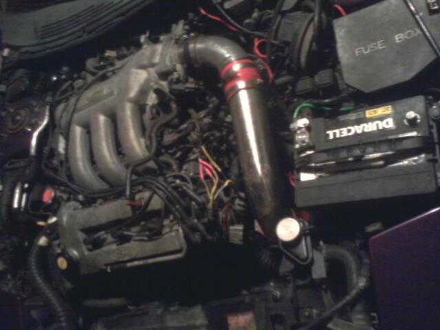 Car engine bay with Duracell battery, fuse box, and silver intake pipe.