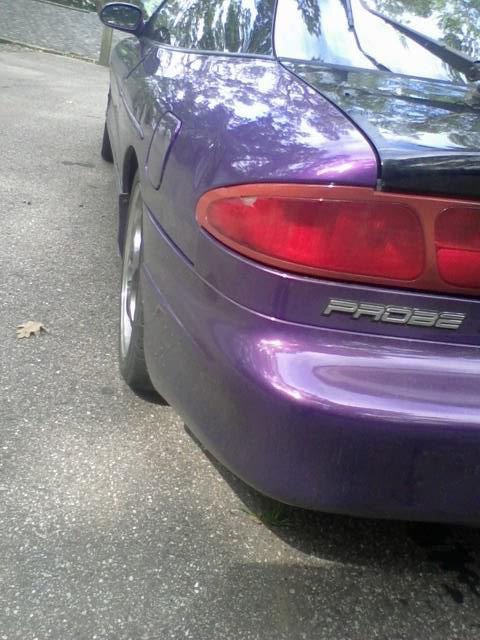 1995 Ford Probe - The rear passenger side quarter of a purple 1995 Ford Probe is shown, parked on an asphalt surface with some gravel and a fallen leaf. The…