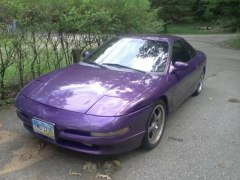 1995 Ford Probe - A purple 1995 Ford Probe is parked on an asphalt driveway surrounded by greenery. The car is viewed from a low angle, showcasing its front…