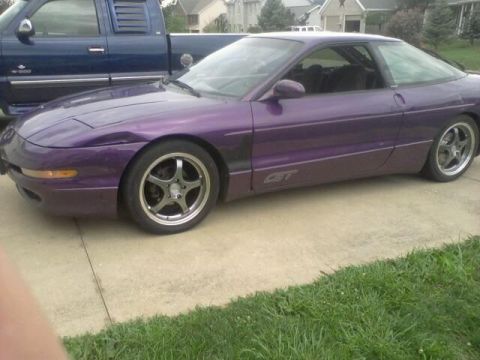 1995 Ford Probe - A purple 1995 Ford Probe GT is parked on a concrete driveway next to a blue pickup truck. The car is viewed from the side with the front wh…