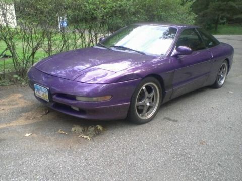 1995 Ford Probe - A purple 1995 Ford Probe is parked on an asphalt driveway with green bushes and trees in the background. The car is viewed from a 3/4 front…