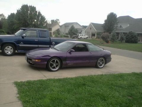 1995 Ford Probe - A purple 1995 Ford Probe GT is parked on a concrete driveway next to a blue pickup truck. The car is viewed from the side, showing its spor…