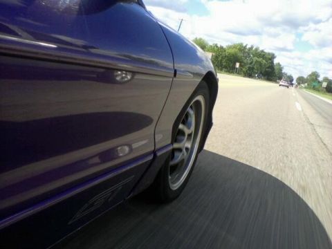 1995 Ford Probe - A low-angle side view of a purple car driving on a road. The car's front wheel and fender are prominent in the foreground, with a shadow ca…