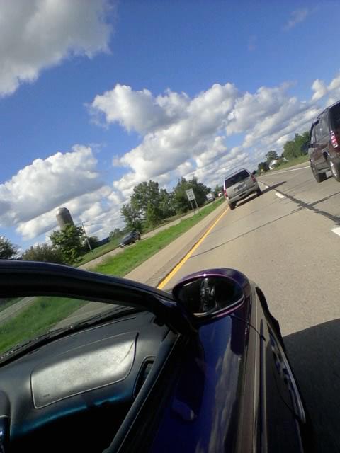 1995 Ford Probe - The driver's side interior and exterior of a dark purple 1995 Ford Probe is visible as it travels on a highway. The car is in motion, with…