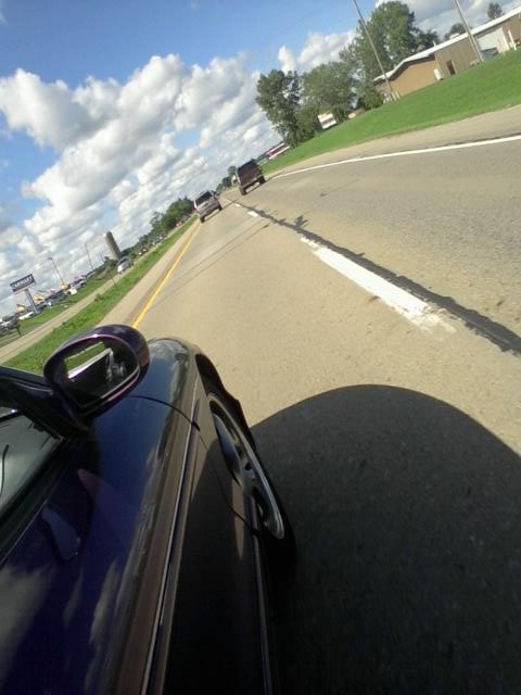 1995 Ford Probe - A low-angle, wide shot of a dark purple coupe driving on a highway. The car's side mirror and part of the front fender are visible in the f…