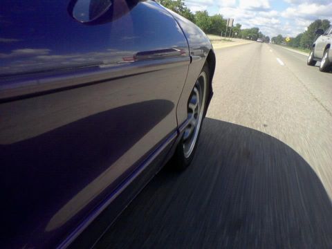 1995 Ford Probe - Side view of a dark purple coupe driving on a highway. The car's reflection of the sky and clouds is visible on its side panel.