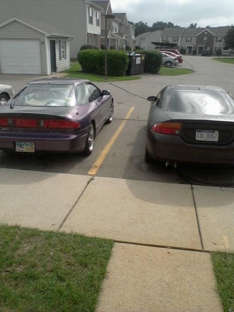 1995 Ford Probe - Two coupes are parked in a residential parking lot. The car on the left is purple and the car on the right is dark gray.