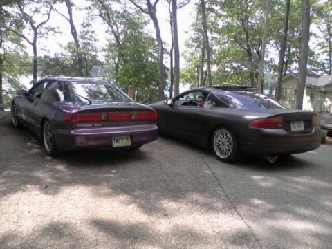 1995 Ford Probe - Two sporty coupes are parked on a paved surface surrounded by trees. The car on the left is purple and the car on the right is dark gray.