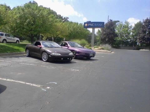 1995 Ford Probe - Two coupes are parked in a paved lot in front of a line of trees and a PNC Bank sign. The car in the foreground is dark gray with silver ri…