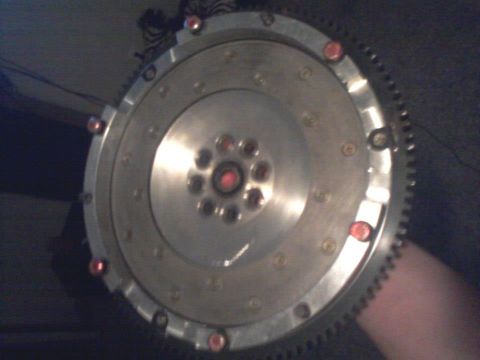 1995 Ford Probe - A close-up, slightly blurry, overhead view of a silver flywheel with a ring of teeth around its outer edge. Red bolts or studs are visible…