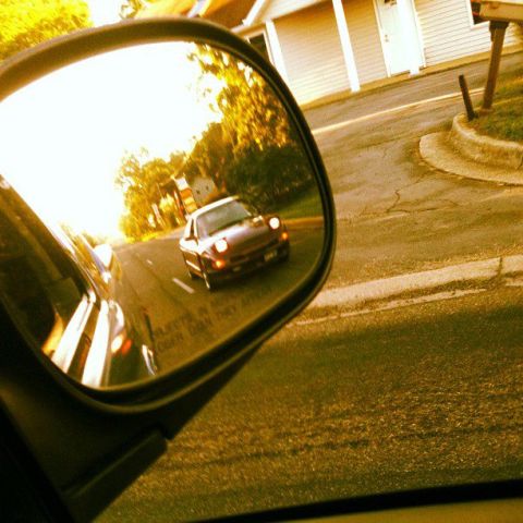 1995 Ford Probe - A dark-colored coupe is reflected in a side mirror, driving down a road with trees and houses visible in the background. The sun is shining…