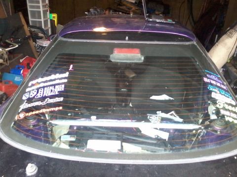 1995 Ford Probe - The rear window of a purple car is covered in various stickers. The car is parked indoors, possibly in a garage, with various items visible…