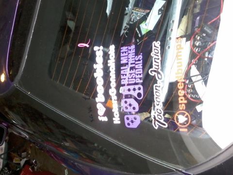 1995 Ford Probe - A close-up, angled shot of the rear window of a dark purple car, featuring several vinyl decals. Visible text includes "i love boobies!", "…