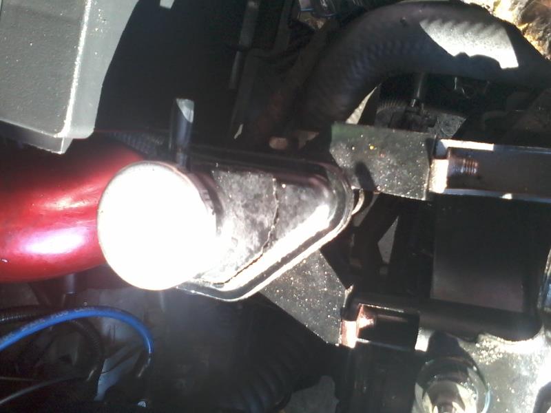 Close-up of a black bracket holding a white-capped cylindrical part in a vehicle engine bay.