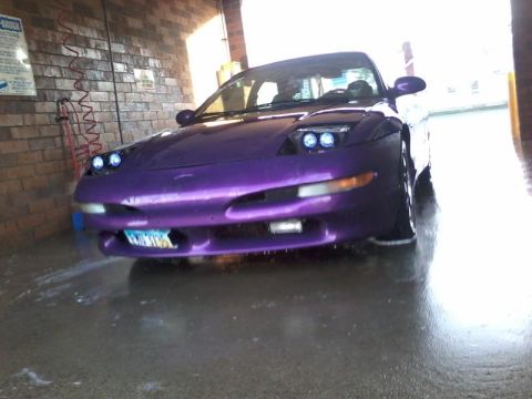 1995 Ford Probe - A low-angle front view of a purple 1995 Ford Probe parked in a car wash bay. The car's headlights are illuminated, and water and soap suds…