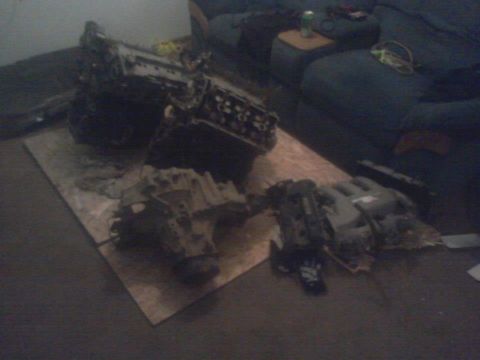 1995 Ford Probe - Two disassembled vehicle engines and transmissions are laid out on a piece of cardboard on the floor. A dark blue couch and a small wooden…