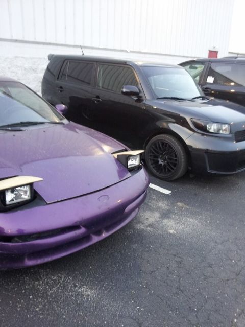 1995 Ford Probe - The front end of a purple sports car with its pop-up headlights partially raised is parked next to a black SUV. The sports car has a damage…
