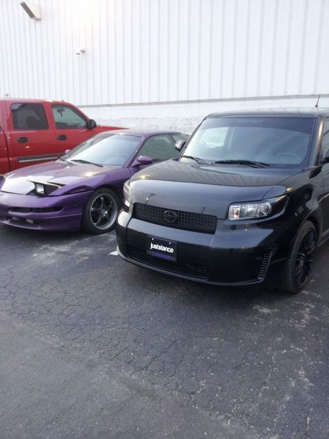 1995 Ford Probe - A purple 1995 Ford Probe with pop-up headlights is parked next to a black Scion xB. A red pickup truck is visible behind the Probe.