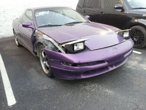 1995 Ford Probe - A damaged purple 1995 Ford Probe is parked on asphalt next to a black SUV. The front fender and hood on the driver's side are missing, reve…