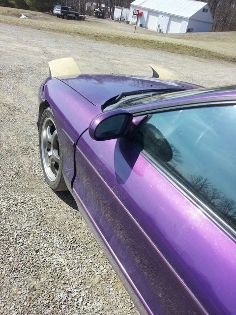 1995 Ford Probe - A close-up, low-angle shot shows the driver's side of a purple 1995 Ford Probe with its pop-up headlights partially raised. The car is park…