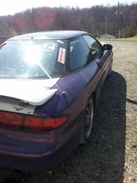 1995 Ford Probe - The rear passenger side of a purple 1990s Ford Probe is shown parked on a gravel and grass surface. A white spoiler is mounted on the trunk…