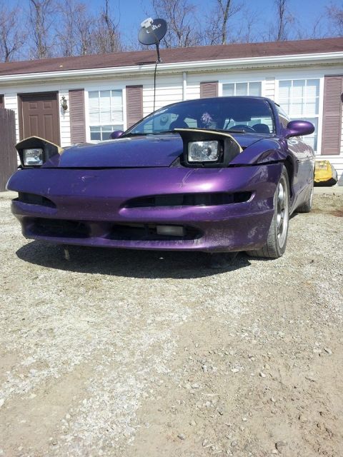 1995 Ford Probe - A front-facing view of a modified purple 1995 Ford Probe with pop-up headlights. The car is parked on a gravel driveway in front of a house.