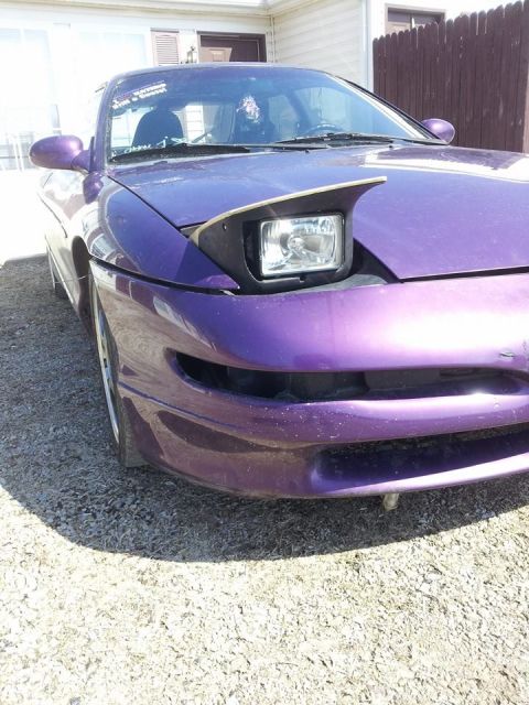 1995 Ford Probe - A low-angle, front-view shot of a purple 1995 Ford Probe with its hood popped open, revealing a pop-up headlight. The car is parked on a gr…