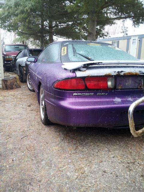 1995 Ford Probe - The rear passenger side of a purple Ford Probe is shown covered in ice. The car is parked outdoors on a gravel surface next to trees and a…