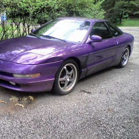 1995 Ford Probe - A purple 1995 Ford Probe is parked on a gravel surface next to a dense green hedge. The car is viewed from a 3/4 front angle, showing its s…