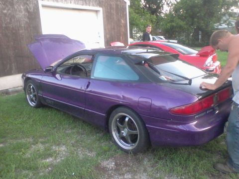 1995 Ford Probe - A purple 1995 Ford Probe with its hood open is parked on grass in front of a wooden building with a garage door. A red car is visible behin…
