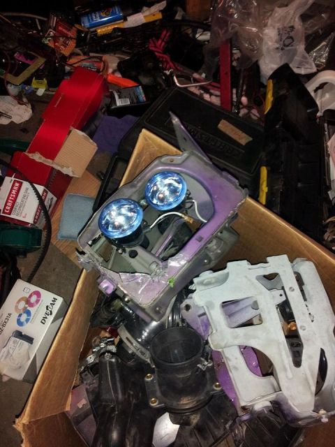 1995 Ford Probe - A close-up view of a disassembled car headlight assembly with two blue projector headlights, resting in a cardboard box amidst other car pa…
