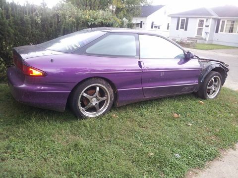 1995 Ford Probe - The side view of a purple, two-door Ford Probe GT is parked on a grassy lawn next to a suburban street. The front fender is missing, reveal…
