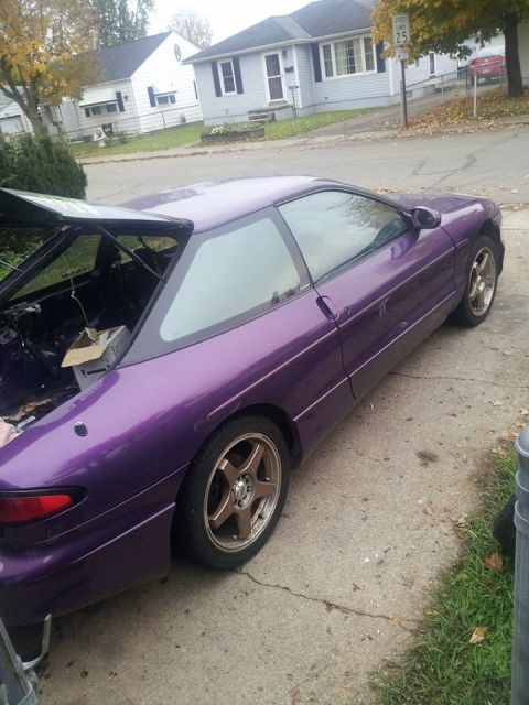 1995 Ford Probe - A purple 1995 Ford Probe is parked on a concrete driveway in front of a residential house. The rear hatch is open, revealing the interior.
