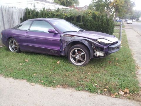 1995 Ford Probe - A purple Ford Probe GT is parked on the grass next to a sidewalk. The front end of the car is damaged and missing body panels and headlight…