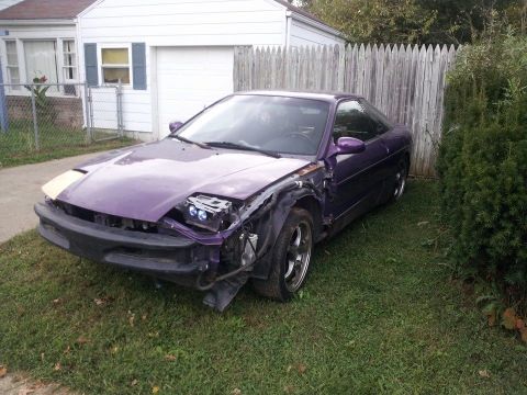1995 Ford Probe - A damaged purple 1995 Ford Probe is parked on a grassy lawn next to a wooden fence and a white house. The front bumper and headlight on the…