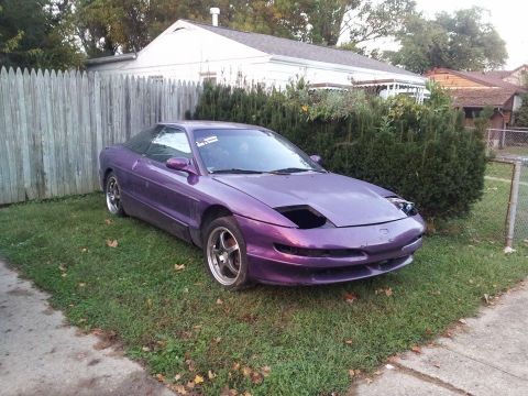 1995 Ford Probe - A purple Ford Probe is parked on a grassy lawn in front of a wooden fence and a white house. The car has its headlights retracted, revealin…
