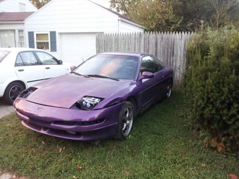 1995 Ford Probe - A purple 1995 Ford Probe is parked on a grassy lawn next to a white sedan and a wooden fence. The Probe's headlights are exposed, and its f…