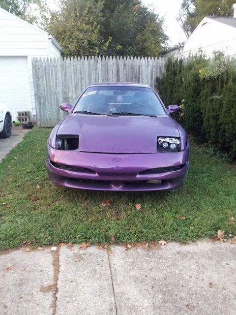 1995 Ford Probe - Front view of a purple 1995 Ford Probe with custom headlights parked on a grassy lawn in front of a wooden fence.