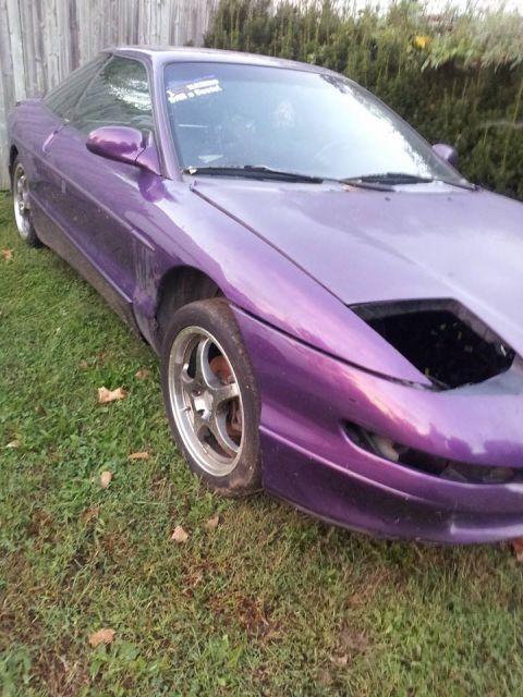 1995 Ford Probe - A purple 1995 Ford Probe is parked on a grassy lawn next to a wooden fence. The front of the car is visible, with the hood open and the hea…