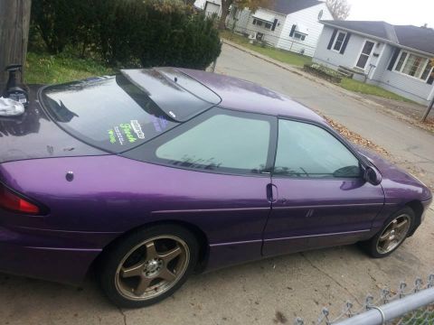 1995 Ford Probe - A purple 1995 Ford Probe is parked on a concrete driveway next to a street with houses in the background. The car has tinted windows and go…
